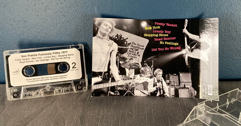 May include: A cassette tape of the Sex Pistols album "Furiously Filthy 1977". The cassette tape is in a clear plastic case. The album cover features a black and white photo of the band performing on stage. The album title is printed in white on a black background. The track list is printed in white on a black background. The album cover also includes the text "The Great South East Music Hall & Emporium Atlanta".