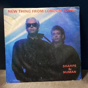 Peut inclure: Une pochette de disque avec un fond bleu représentant deux hommes en vestes noires. Le texte sur la pochette est "New Thing From London Town" et "Sharpe & Numan".