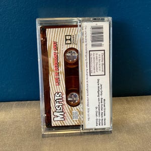 May include: A cassette tape with a brown and gold label. The label features the band name "Misfits" in large white letters and the text "Live in Germany 1997". The cassette tape is in a clear plastic case.