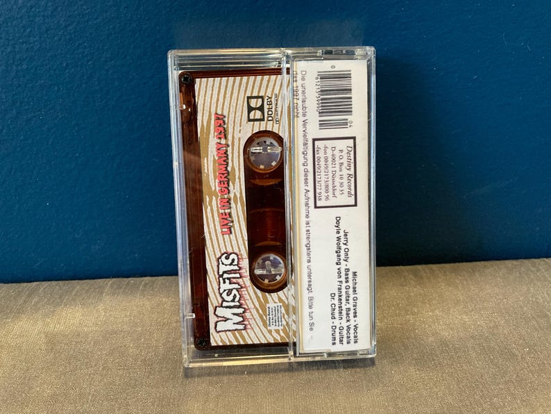 May include: A cassette tape with a brown and gold label. The label features the band name "Misfits" in large white letters and the text "Live in Germany 1997". The cassette tape is in a clear plastic case.