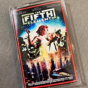 Puede incluir: Una caja de casete de plástico transparente que contiene la película El quinto elemento. La portada muestra a Milla Jovovich, Bruce Willis y Gary Oldman con un horizonte urbano futurista, vehículos voladores y el título en letras blancas.