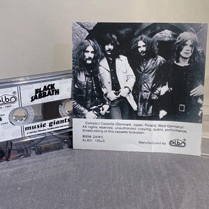 K&ouml;nnte beinhalten: Eine transparente Kassette mit dem Bandnamen "Black Sabbath" und den Worten "music giants". Ein Schwarz-Wei&szlig;-Foto der Band befindet sich neben der Kassette. Die Kassette ist mit "elbo" und "BIEM ZAIKS" beschriftet.