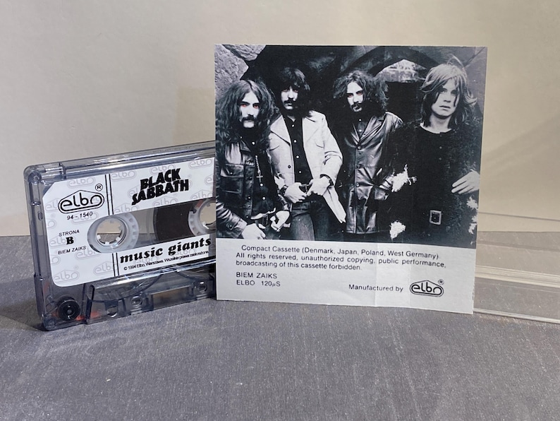 K&ouml;nnte beinhalten: Eine transparente Kassette mit dem Bandnamen "Black Sabbath" und den Worten "music giants". Ein Schwarz-Wei&szlig;-Foto der Band befindet sich neben der Kassette. Die Kassette ist mit "elbo" und "BIEM ZAIKS" beschriftet.