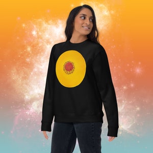 Puede incluir: Una sudadera negra con un diseño circular amarillo con un centro rojo y dorado. La sudadera es una prenda informal y cómoda.