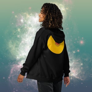 Puede incluir: Una sudadera con capucha negra con un diseño circular amarillo en la espalda. El círculo tiene un patrón rojo y dorado en su interior.