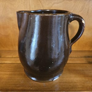 Puede incluir: Una jarra de cer&aacute;mica marr&oacute;n oscuro con un cuerpo redondeado y un asa curva. La jarra tiene un acabado brillante y un peque&ntilde;o pico para verter. La pieza de cer&aacute;mica hecha a mano es adecuada para servir bebidas.