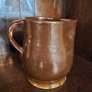 Puede incluir: Una taza de cer&aacute;mica marr&oacute;n con base redondeada y asa curva. La taza tiene un acabado brillante y parece hecha a mano. La taza est&aacute; sobre una superficie de madera.