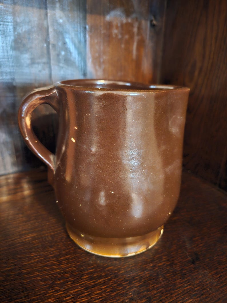 Puede incluir: Una taza de cer&aacute;mica marr&oacute;n con base redondeada y asa curva. La taza tiene un acabado brillante y parece hecha a mano. La taza est&aacute; sobre una superficie de madera.