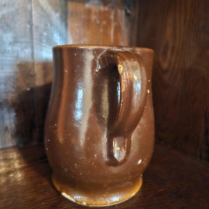 Puede incluir: Una taza de cer&aacute;mica marr&oacute;n con un asa curva. La taza tiene un acabado brillante y una forma ligeramente irregular. La taza est&aacute; sobre una superficie de madera. La taza es de color marr&oacute;n oscuro.