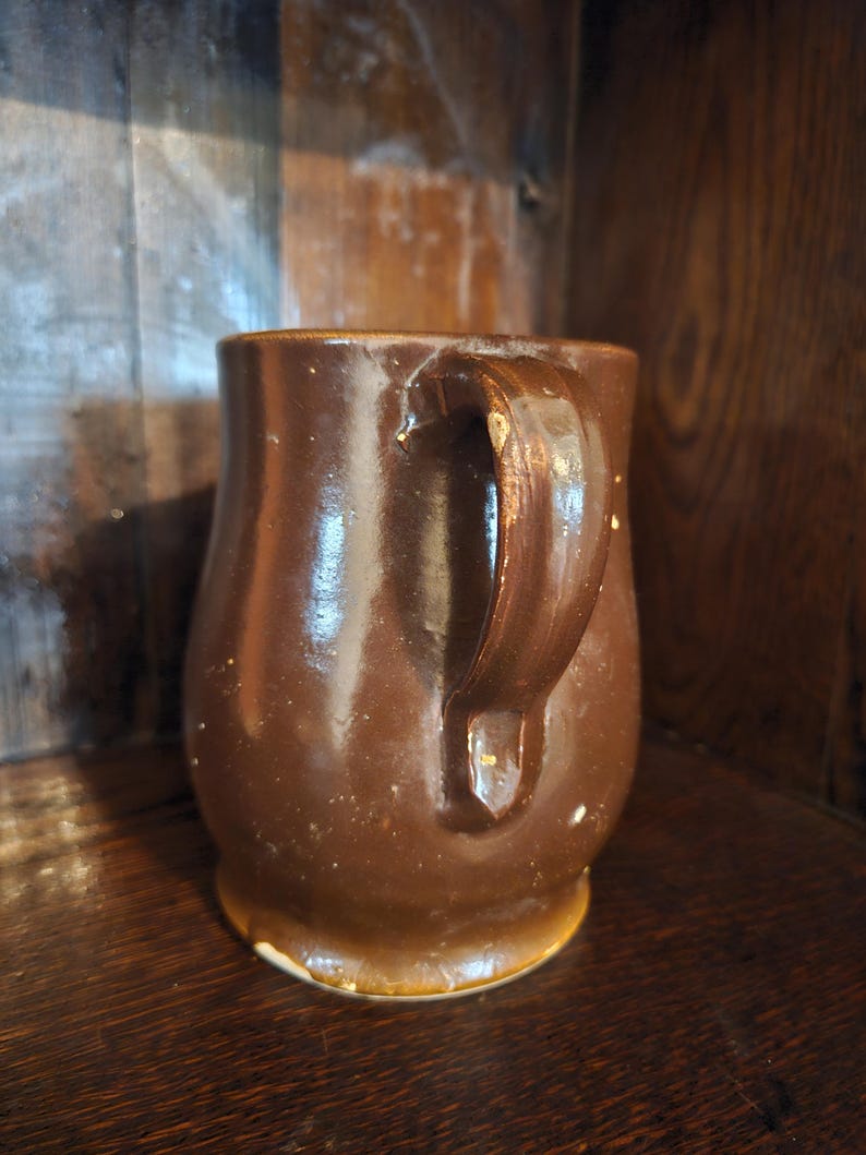 Puede incluir: Una taza de cer&aacute;mica marr&oacute;n con un asa curva. La taza tiene un acabado brillante y una forma ligeramente irregular. La taza est&aacute; sobre una superficie de madera. La taza es de color marr&oacute;n oscuro.