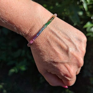 May include: A delicate beaded bracelet featuring a gradient of small, polished gemstones in a spectrum of colors including pink, purple, blue, green, and yellow. The beads are strung in a single row, creating a vibrant, multi-colored rainbow effect.