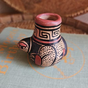 May include: A small, hand-painted ceramic bird figurine. The bird is primarily black with tan and red accents. It features geometric patterns and a stylized design, with a round body and a small spout. The figurine is likely a decorative item.