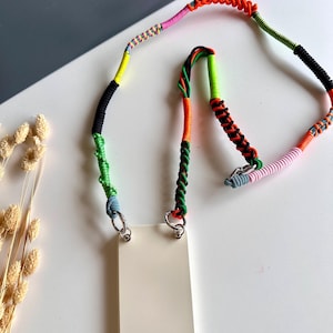 Könnte beinhalten: Eine cremefarbene Smartphone-Hülle mit einem mehrfarbigen, handgewebten Kordelband. Das Band weist verschiedene Texturen und Muster in Neonorange, Hellgrün, Pink, Schwarz und Blau auf und ist mit zwei silbernen Metallringen an der Hülle befestigt.