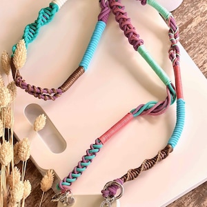Op de afbeelding: Een kleurrijke macramé telefoonlanyard met een verscheidenheid aan kleuren, waaronder turquoise, roze, paars, bruin en wit. De lanyard heeft een gevlochten ontwerp en metalen hardware met bedels. De lanyard is ontworpen om een telefoon vast te houden.