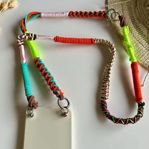 Könnte beinhalten: Ein Telefonband mit einem mehrfarbigen, geflochtenen Kordeldesign, das Abschnitte aus neon-grünem, leuchtend orangefarbenem, türkisfarbenem, hellrosa und braunem Garn aufweist. Das Band ist mit zwei silberfarbenen Metallringen an einer cremeweißen Handyhülle befestigt.