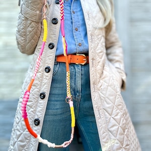Puede incluir: Una correa para perros colorida con un diseño trenzado. La correa presenta una combinación de cuerda rosa, amarilla, naranja y blanca. La correa está sujeta a un anillo de metal dorado. La persona lleva una chaqueta acolchada beige, vaqueros azules y un cinturón naranja.