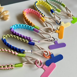Könnte beinhalten: Eine Sammlung von fünf geflochtenen Paracord-Handschlaufen mit mehrfarbigen Mustern. Jedes Schlüsselband enthält eine beigefarbene Kordelschlaufe, einen kleinen Metallring und einen T-förmigen Kunststoffaufsatz in verschiedenen Farben wie Pink, Blau, Lila, Orange und Gelb.