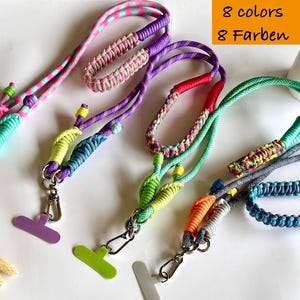 Op de afbeelding: Een verzameling kleurrijke telefoonlanyards in verschillende tinten, waaronder roze, paars, groen en blauw. Elke lanyard heeft een gevlochten of geweven ontwerp en een metalen sluiting om aan een telefoon te bevestigen. De afbeelding bevat de tekst "8 kleuren".