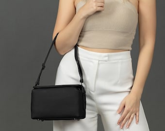 Può includere: Una borsa a tracolla in pelle nera con chiusura a lembo. La borsa è indossata da una persona che indossa un top beige halter e pantaloni bianchi.