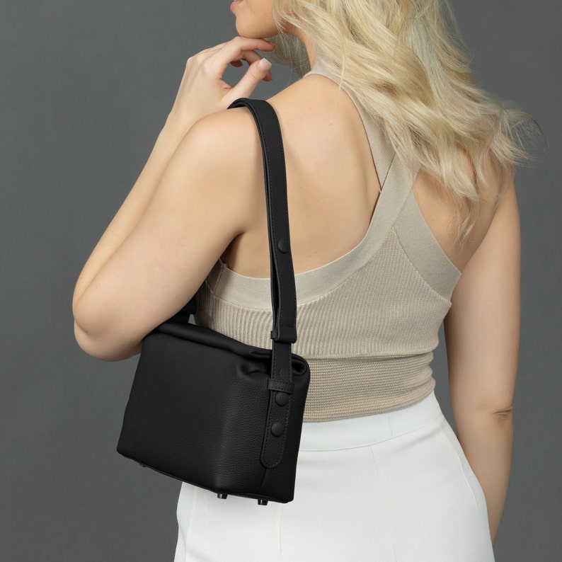 May include: A black leather shoulder bag with a flap closure and a long adjustable strap. The bag has a boxy shape and a textured finish.