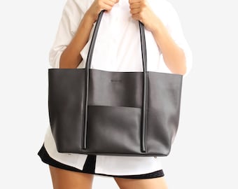 May include: A black tote bag with long handles and an exterior pocket. The bag is made of a smooth material and has a minimalist design. The bag is held by a person wearing a white shirt.