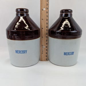 May include: Two antique stoneware jugs. The jugs have a light gray base and a dark brown, glossy top. The word "MERCURY" is printed in blue on the front of each jug. A wooden ruler is placed between the jugs, measuring approximately 8 inches in height.