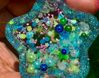 May include: A star-shaped resin shaker charm featuring a translucent teal border filled with iridescent glitter. The center contains a mix of loose elements including small blue and green spherical beads, holographic star-shaped sequins, and various geometric shapes.