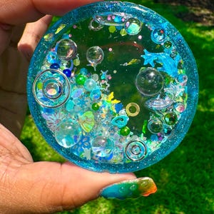 Pode incluir: Um porta-copos circular de resina com uma base azul-petr&oacute;leo transl&uacute;cida, preenchido com v&aacute;rios elementos decorativos incrustados, incluindo esferas de vidro transparente, pingentes em espiral prateados, lantejoulas em forma de estrela e contas verdes.