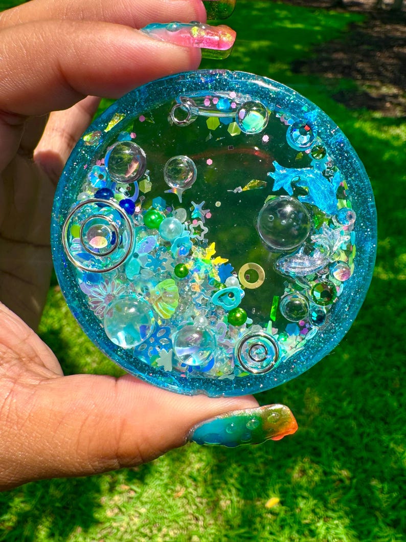 Pode incluir: Um porta-copos circular de resina com uma base azul-petr&oacute;leo transl&uacute;cida, preenchido com v&aacute;rios elementos decorativos incrustados, incluindo esferas de vidro transparente, pingentes em espiral prateados, lantejoulas em forma de estrela e contas verdes.