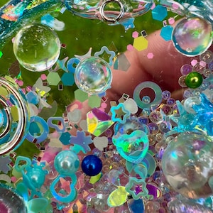 Pode incluir: Um pingente circular tipo shaker de resina transparente preenchido com uma variedade de enfeites coloridos, incluindo esferas iridescentes, contas azuis e verdes, formas hologr&aacute;ficas de estrelas e cora&ccedil;&otilde;es e pequenas lantejoulas geom&eacute;tricas.