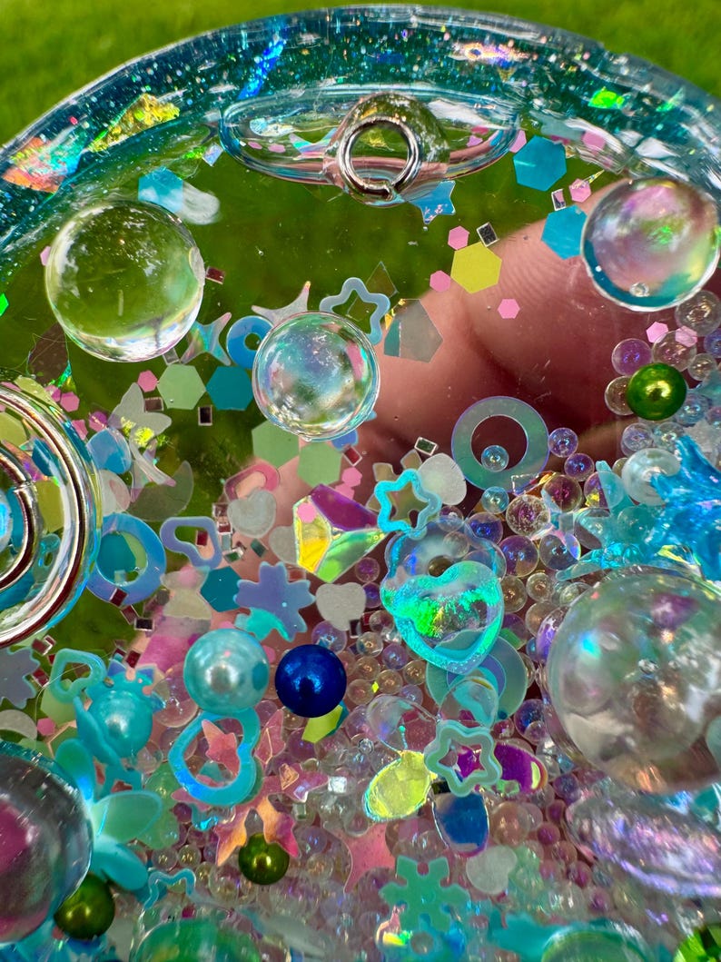 Pode incluir: Um pingente circular tipo shaker de resina transparente preenchido com uma variedade de enfeites coloridos, incluindo esferas iridescentes, contas azuis e verdes, formas hologr&aacute;ficas de estrelas e cora&ccedil;&otilde;es e pequenas lantejoulas geom&eacute;tricas.