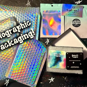 Pode incluir: Uma cole&ccedil;&atilde;o de envelopes almofadados hologr&aacute;ficos, uma bolsa verde-menta, um cart&atilde;o de agradecimento e uma caixa de joias preta contendo dois cart&otilde;es quadrados com o texto Galactic Lagoon. O texto Holographic Packaging est&aacute; em branco.