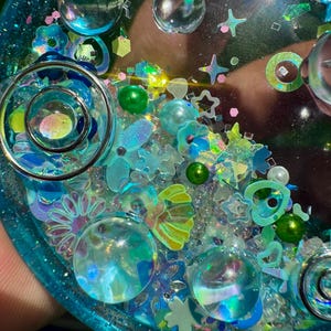 Pode incluir: Uma bandeja circular de resina em tons de azul e verde-azulado, preenchida com v&aacute;rios elementos decorativos embutidos, incluindo esferas de vidro transparente, estrelas iridescentes, pequenas p&eacute;rolas verdes, lantejoulas de flores e cora&ccedil;&otilde;es.
