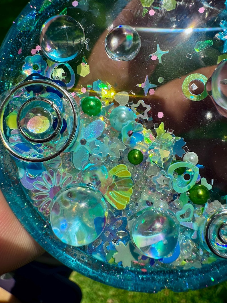 Pode incluir: Uma bandeja circular de resina em tons de azul e verde-azulado, preenchida com v&aacute;rios elementos decorativos embutidos, incluindo esferas de vidro transparente, estrelas iridescentes, pequenas p&eacute;rolas verdes, lantejoulas de flores e cora&ccedil;&otilde;es.