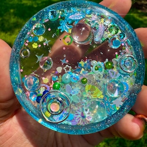 Pode incluir: Um porta-copos circular de resina em tons de azul transl&uacute;cido, com elementos decorativos embutidos, incluindo esferas transparentes, formas de estrelas e flores iridescentes, pequenas contas verdes e an&eacute;is met&aacute;licos, dentro de uma borda azul brilhante.