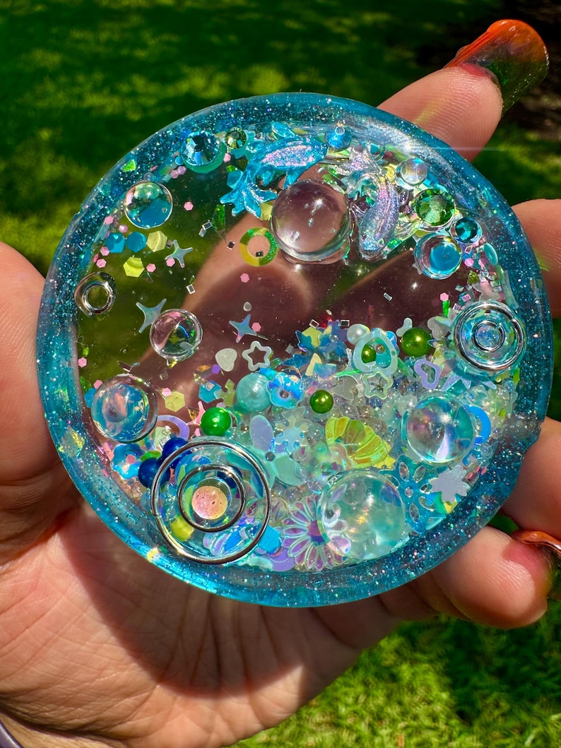 Pode incluir: Um porta-copos circular de resina em tons de azul transl&uacute;cido, com elementos decorativos embutidos, incluindo esferas transparentes, formas de estrelas e flores iridescentes, pequenas contas verdes e an&eacute;is met&aacute;licos, dentro de uma borda azul brilhante.