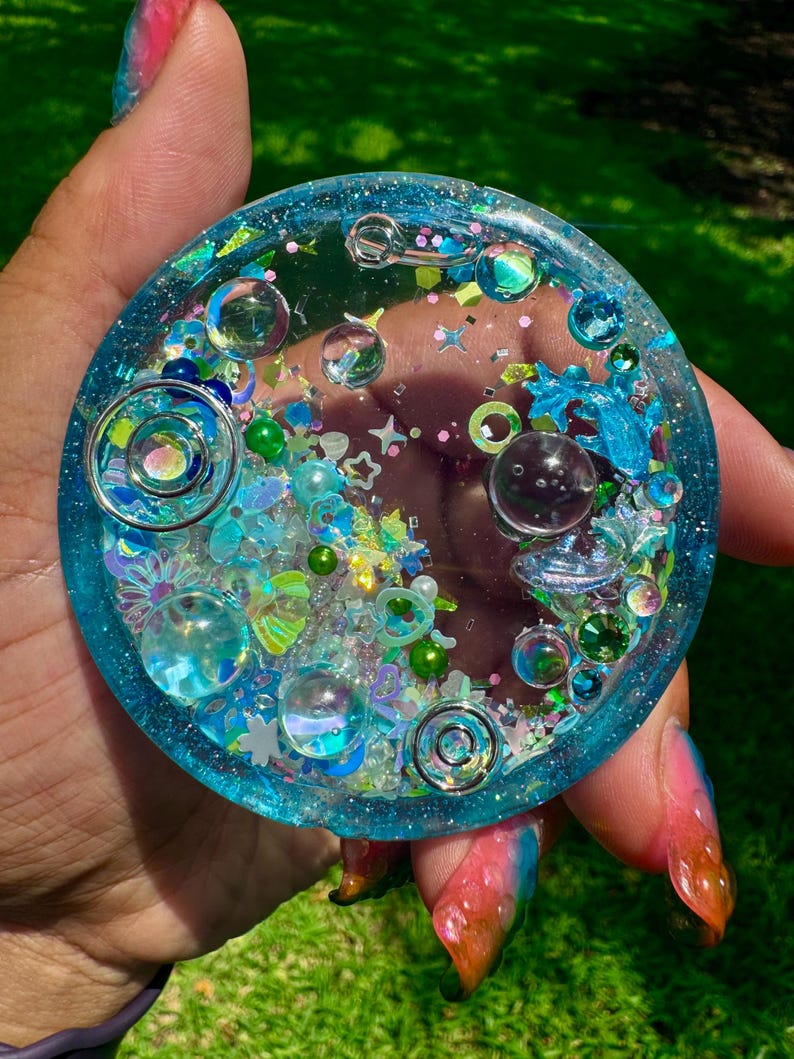 Pode incluir: Um porta-copos circular de resina com uma borda azul transl&uacute;cida preenchida com v&aacute;rios elementos decorativos incrustados, incluindo esferas de vidro transparente, espirais de metal prateado, lantejoulas em forma de estrela e pequenas contas verdes.