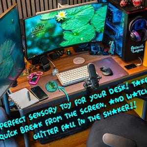 Pode incluir: Um brinquedo sensorial redondo, verde e cheio de glitter sobre uma mesa de madeira. A mesa inclui um computador com dois monitores, teclado, rato, microfone, smartphone, caderno e uma garrafa de &aacute;gua amarela. O texto diz: PERFECT SENSORY TOY FOR YOUR DESK! TAKE A QUICK BREAK FROM THE SCREEN, AND WATCH THE GLITTER FALL IN THE SHAKER!