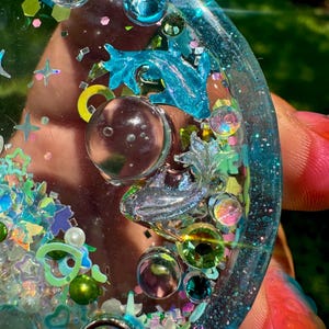 Pode incluir: Um porta-copos circular de resina com um tom azul transl&uacute;cido, incrustado com glitter iridescente, pequenas formas de estrelas e cora&ccedil;&otilde;es, esferas transparentes e duas figuras de golfinhos met&aacute;licos. A superf&iacute;cie &eacute; brilhante.