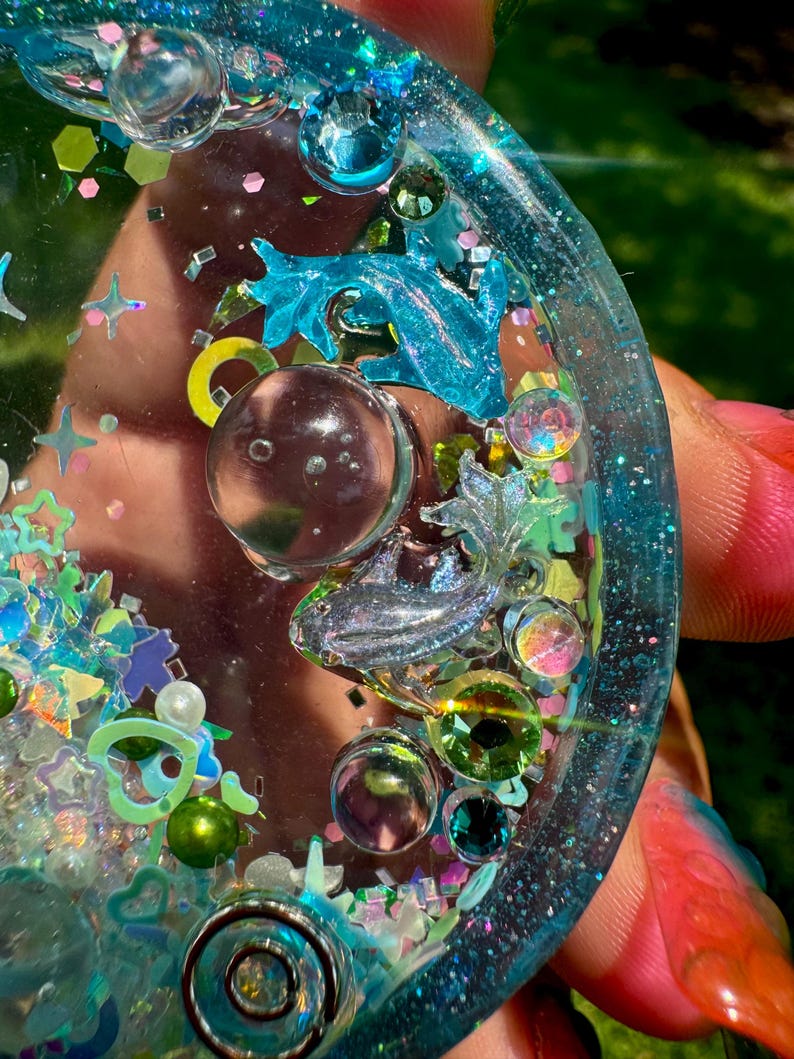 Pode incluir: Um porta-copos circular de resina com um tom azul transl&uacute;cido, incrustado com glitter iridescente, pequenas formas de estrelas e cora&ccedil;&otilde;es, esferas transparentes e duas figuras de golfinhos met&aacute;licos. A superf&iacute;cie &eacute; brilhante.