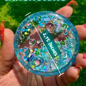 Pode incluir: Um porta-copos circular de resina azul preenchido com v&aacute;rios brilhos, pequenas contas e formas decorativas. O texto "DIMENSIONS" aparece no topo e "2.75 INCHES" est&aacute; impresso no centro com uma linha indicando o di&acirc;metro. "Galactic Lagoon" est&aacute; escrito na parte inferior.
