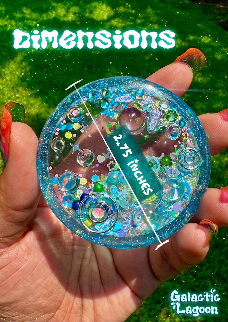 Pode incluir: Um porta-copos circular de resina azul preenchido com v&aacute;rios brilhos, pequenas contas e formas decorativas. O texto "DIMENSIONS" aparece no topo e "2.75 INCHES" est&aacute; impresso no centro com uma linha indicando o di&acirc;metro. "Galactic Lagoon" est&aacute; escrito na parte inferior.