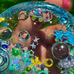 May include: A circular resin tray featuring a translucent blue base embedded with various decorative elements including clear glass spheres, iridescent star-shaped sequins, small green and blue pearls, and assorted metallic glitter pieces!