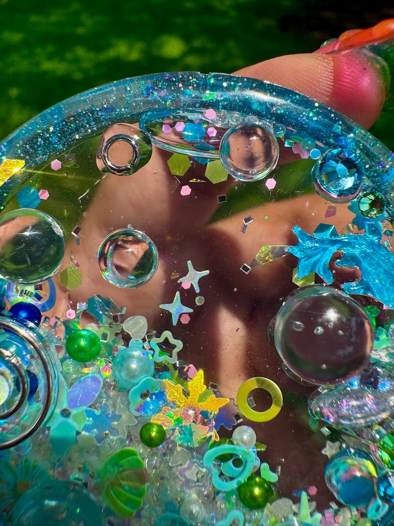 May include: A circular resin tray featuring a translucent blue base embedded with various decorative elements including clear glass spheres, iridescent star-shaped sequins, small green and blue pearls, and assorted metallic glitter pieces!
