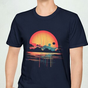 May include: A navy blue t-shirt with a graphic of a sunset over a body of water with a large orange sun and two smaller planets. The graphic is in a retro style with a pixelated look.