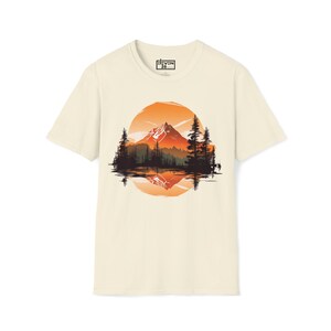 以下が含まれることがあります： クリーム色のTシャツ。山岳風景のデザインが特徴です。グラフィックは、山脈、木々、湖を描き、オレンジ色の夕焼けを背景にしています。シャツはクルーネックと半袖です。ブランドロゴは左上にあります。