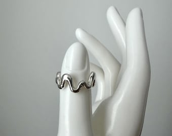 May include: A silver-colored ring with a wavy design. The ring is worn on a white hand. The ring's design is a continuous, flowing wave pattern. The ring is a simple yet elegant piece of jewelry.