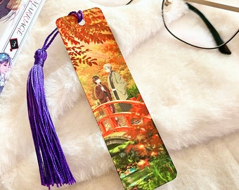 May include: A colorful bookmark featuring an anime-style illustration of two figures on a red bridge in a natural setting. The bookmark has a purple tassel attached. A book and glasses are in the background.