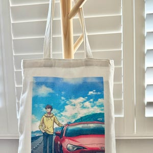 May include: A white canvas tote bag with a printed image of an anime character standing next to a red sports car. The background is a blue sky with white clouds. The car has a Japanese license plate that reads "6-91".