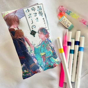 May include: A white canvas pouch with a colorful anime illustration and Japanese text. The text reads "桜降る夜に幸せを" which translates to "Happiness in the night of cherry blossoms falling". The pouch is lying on a white surface with several colorful markers.